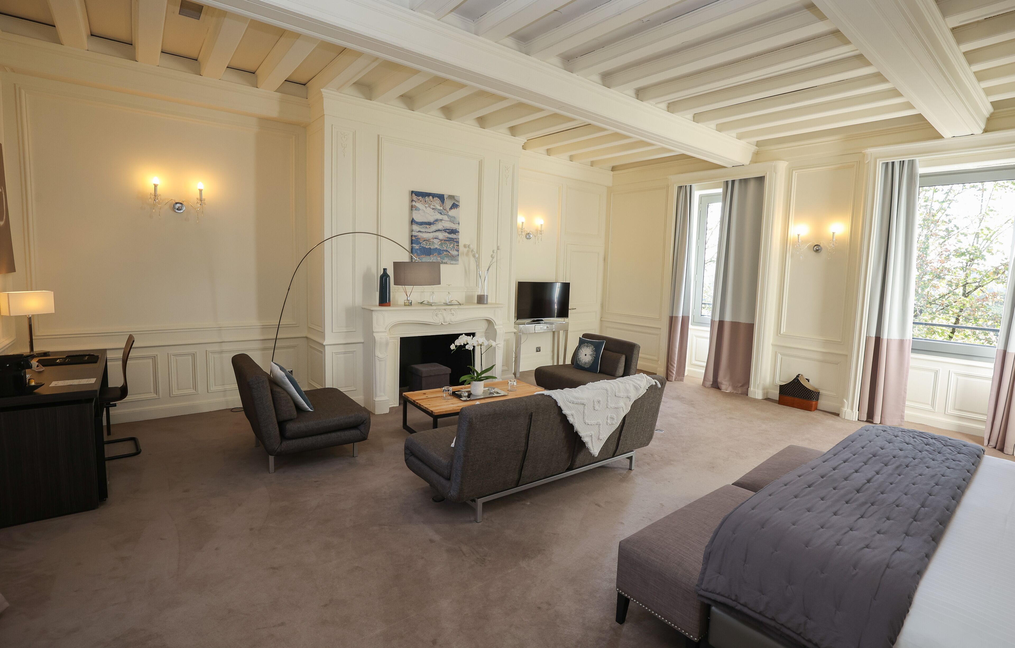 Une chambre d’hôtel avec un canapé, un fauteuil et un lit. On y trouve une cheminée, un téléviseur et un bureau avec une lampe.