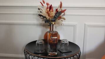Una mesa con un jarrón de flores secas y una botella, junto a dos vasos sobre una mesa de alambre negro frente a una pared blanca.