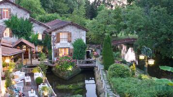 Un jardín junto al río con una casa de piedra, un puente de madera y una zona de estar al aire libre.