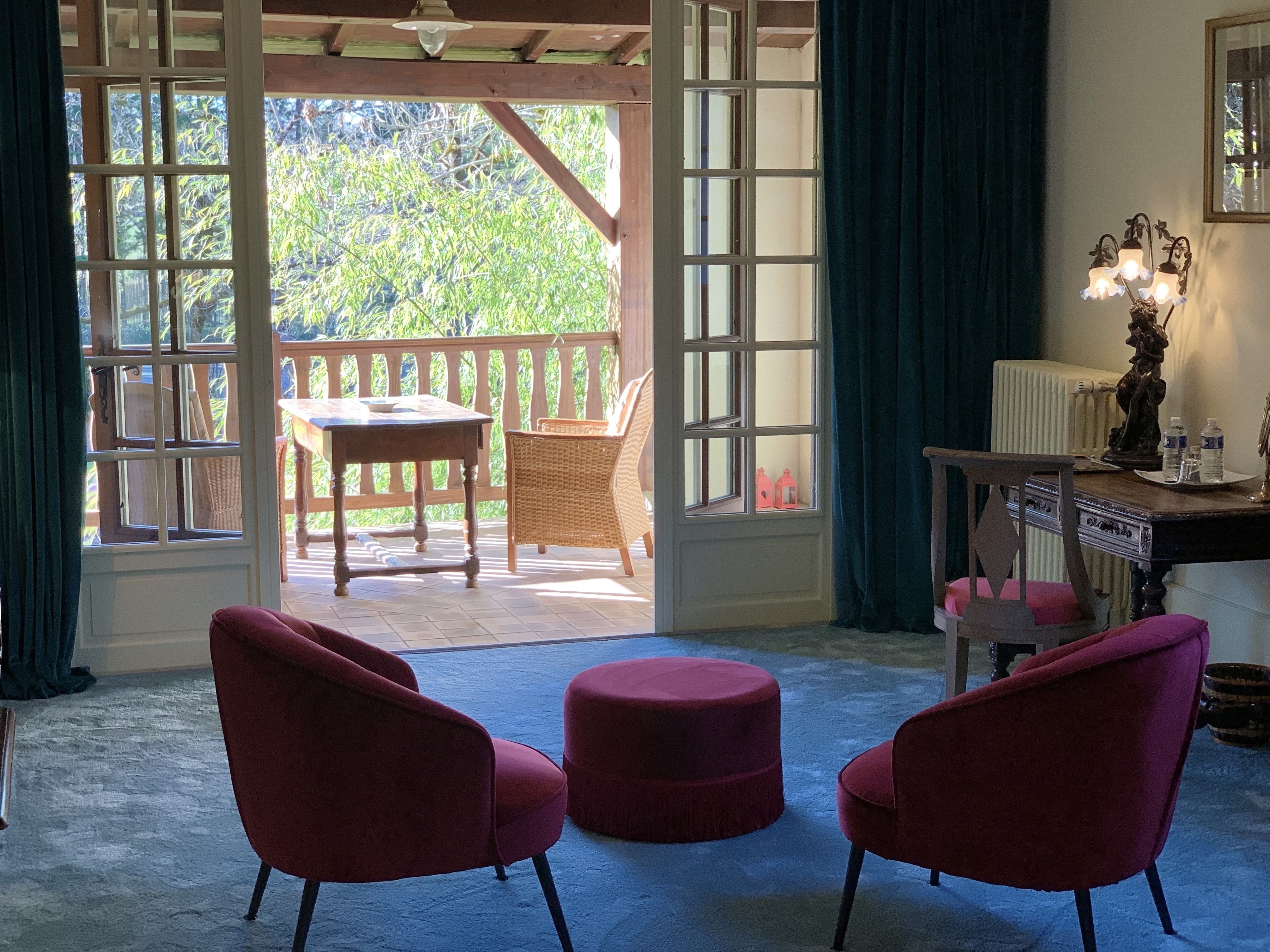 A room with a view of a wooden deck and greenery, two red chairs, a table, and a lamp on a side table.