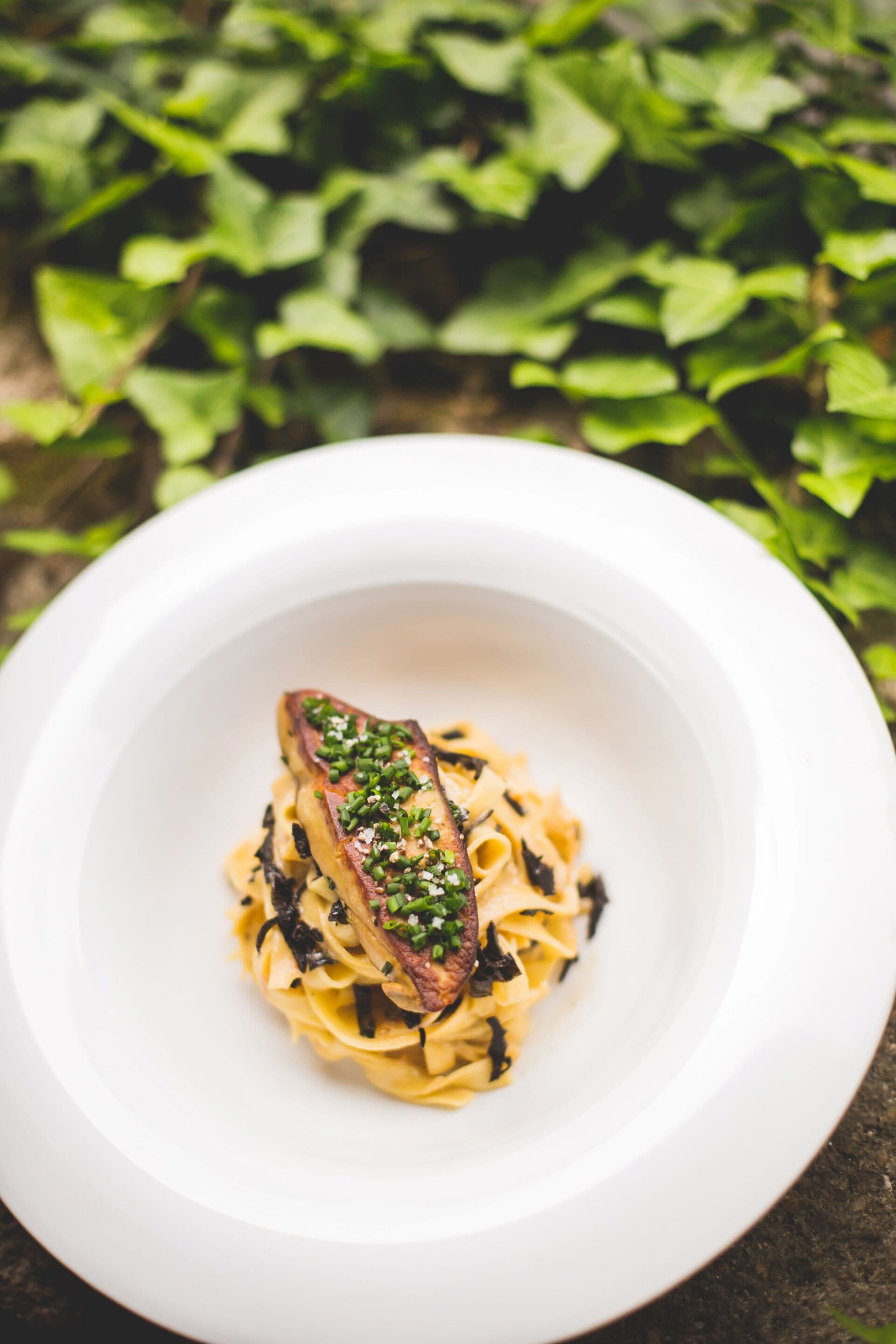 Un tazón blanco de pasta con un trozo de carne y decoración verde sobre una mesa.
