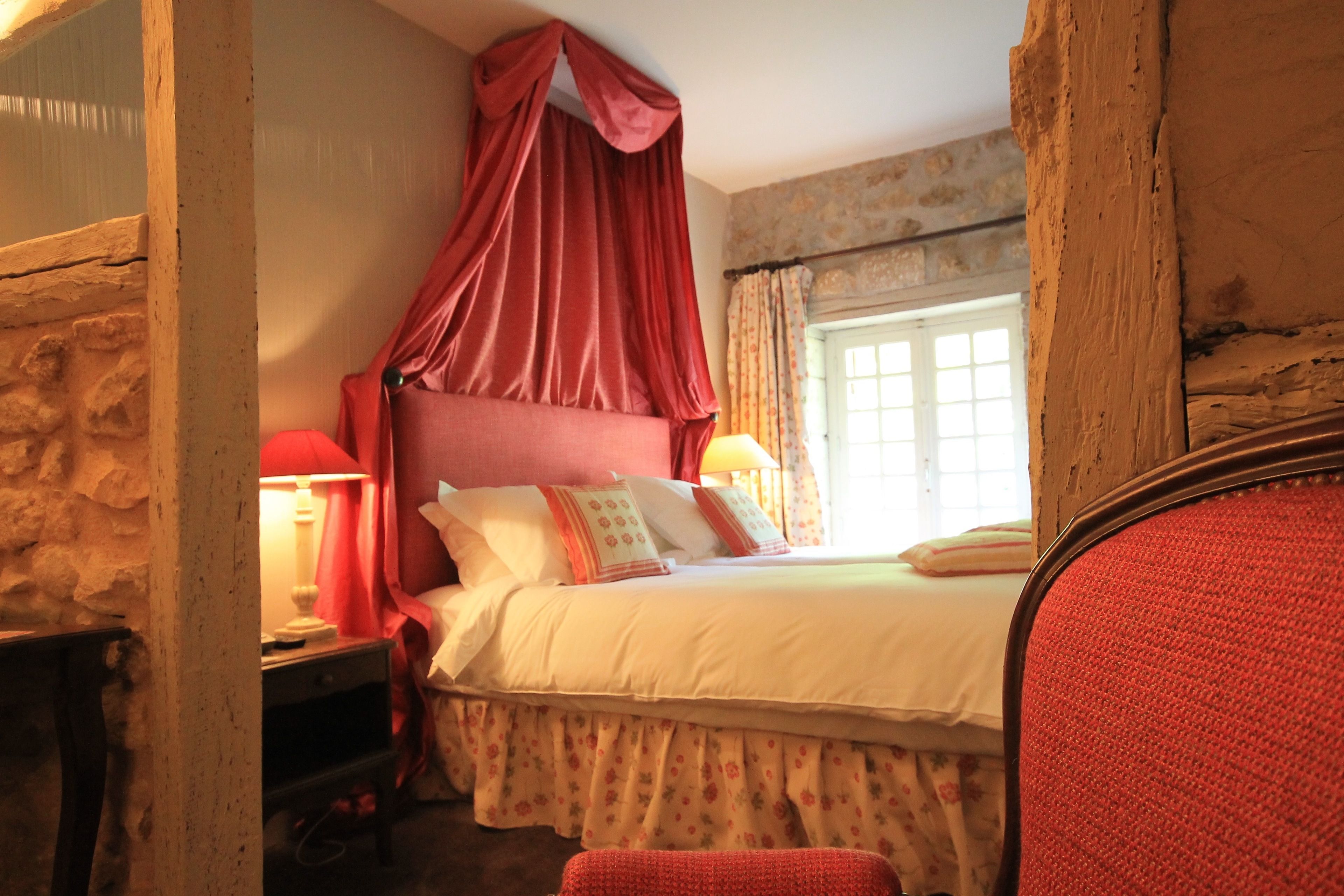 A bedroom with a canopy bed, a nightstand, a lamp, and a window with curtains.
