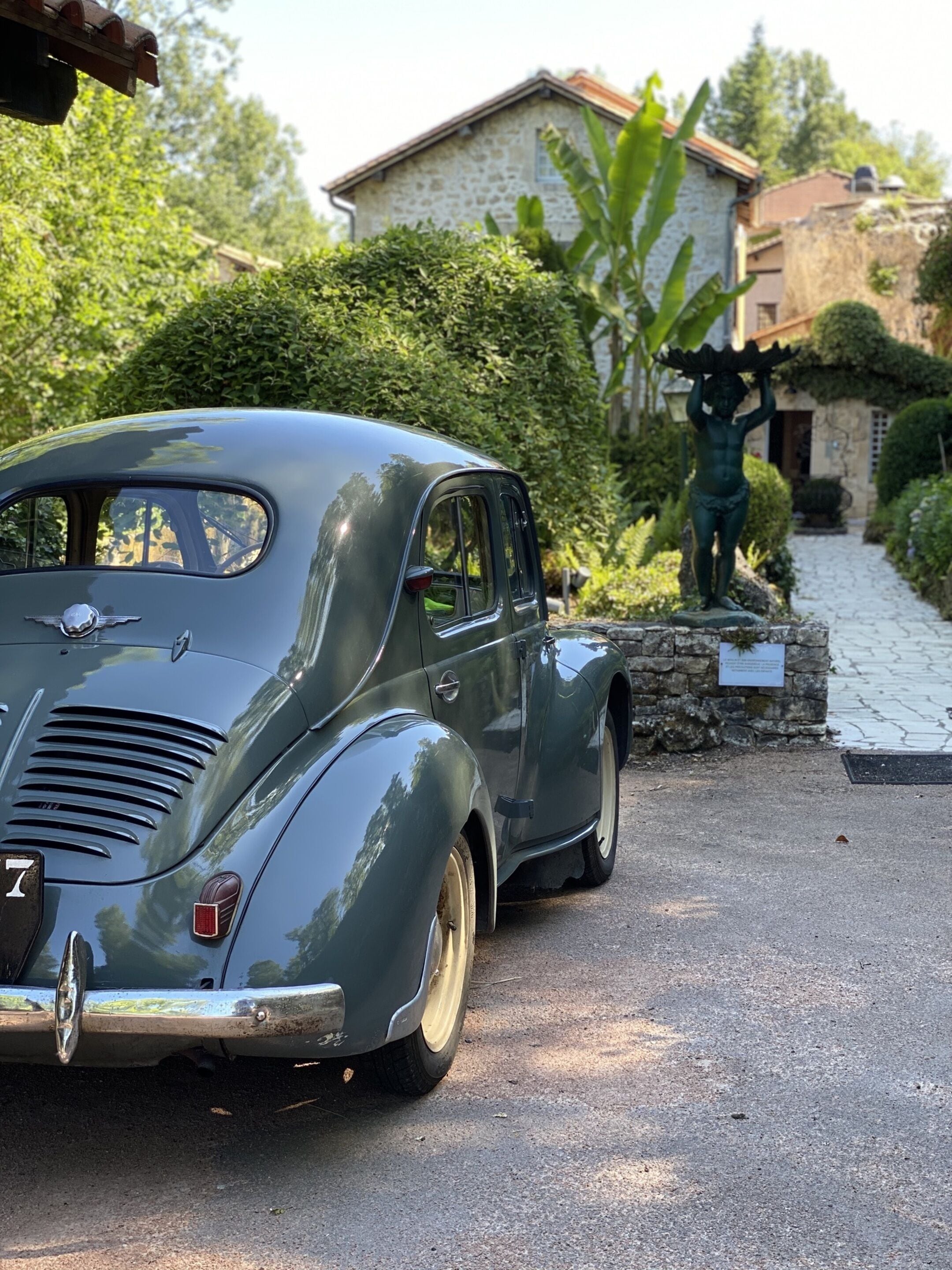 Une voiture ancienne est garée dans une allée bordée d’un mur de pierres, avec une statue en arrière-plan.
