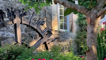 Une construction en pierre dotée d’une grande roue à eau, entourée d’une végétation luxuriante et de fleurs d’un rose éclatant.