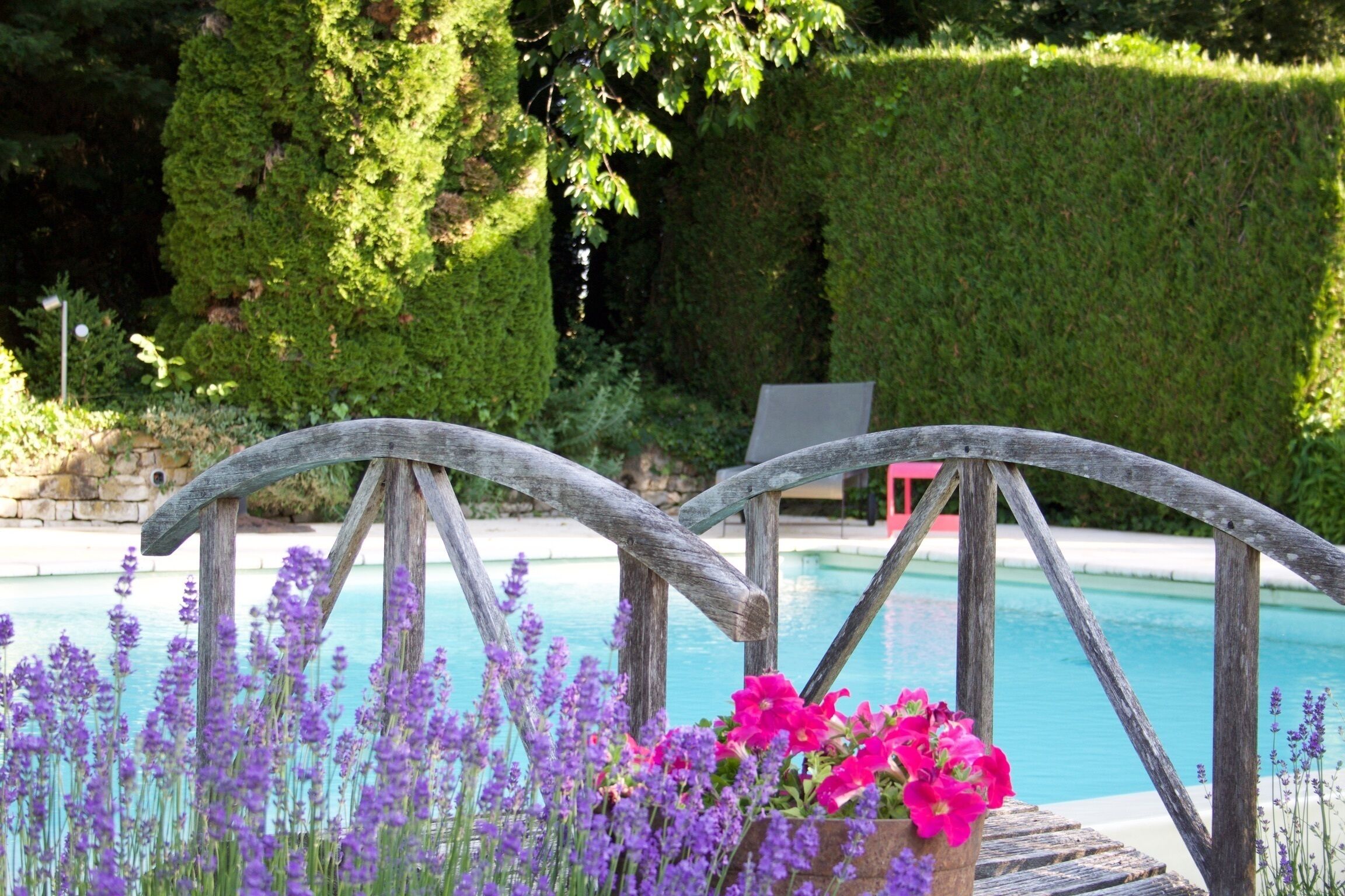 A wooden deck with a pool in the background, surrounded by lush greenery and purple flowers.