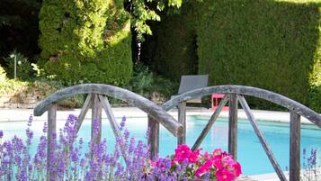 A wooden deck with a pool in the background, surrounded by lush greenery and purple flowers.