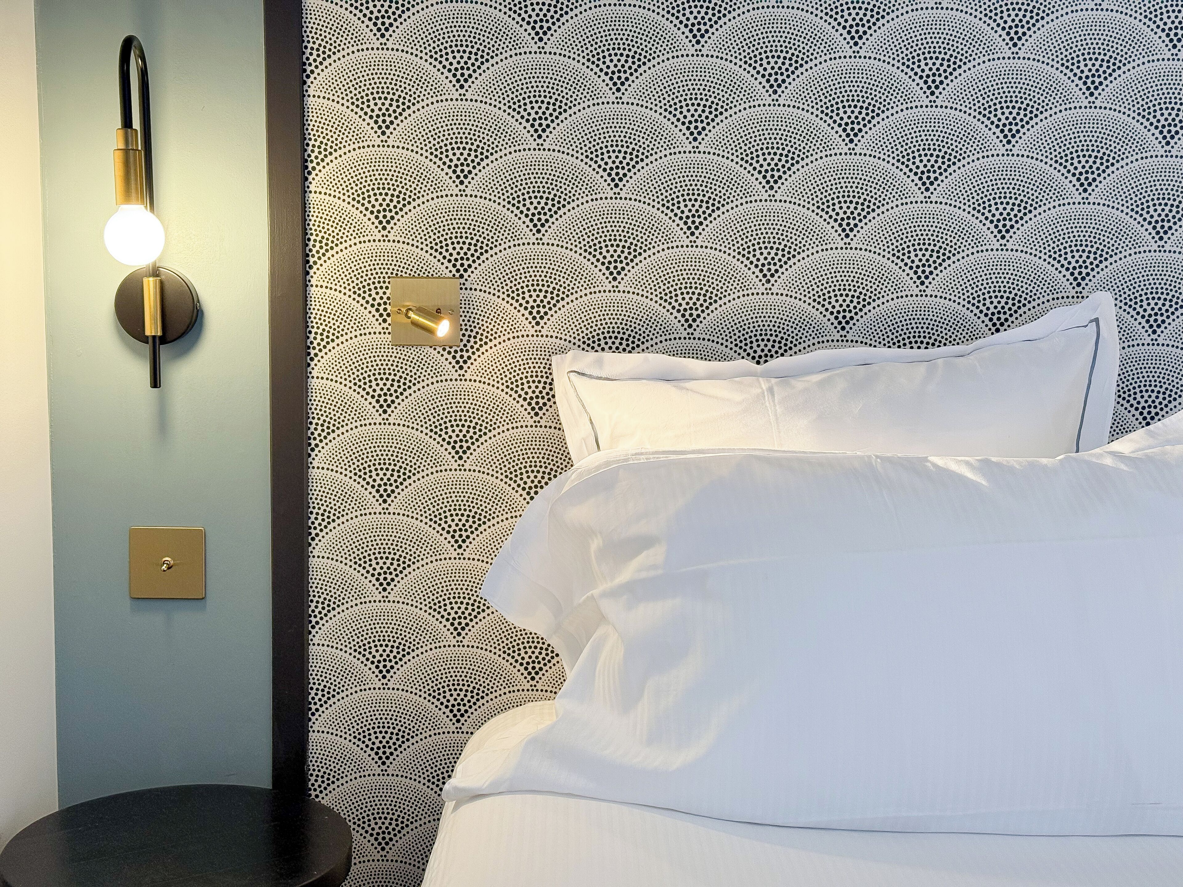 Een bed met witte lakens, een wandlamp en behang met een patroon.