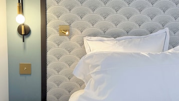 Uma cama com lençóis brancos, luminária fixada na parede e papel de parede estampado.