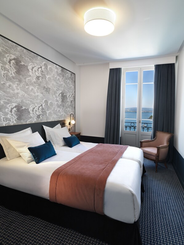 A hotel room with a large bed, a view of the sea, a chair, and a wall with a cloud pattern.