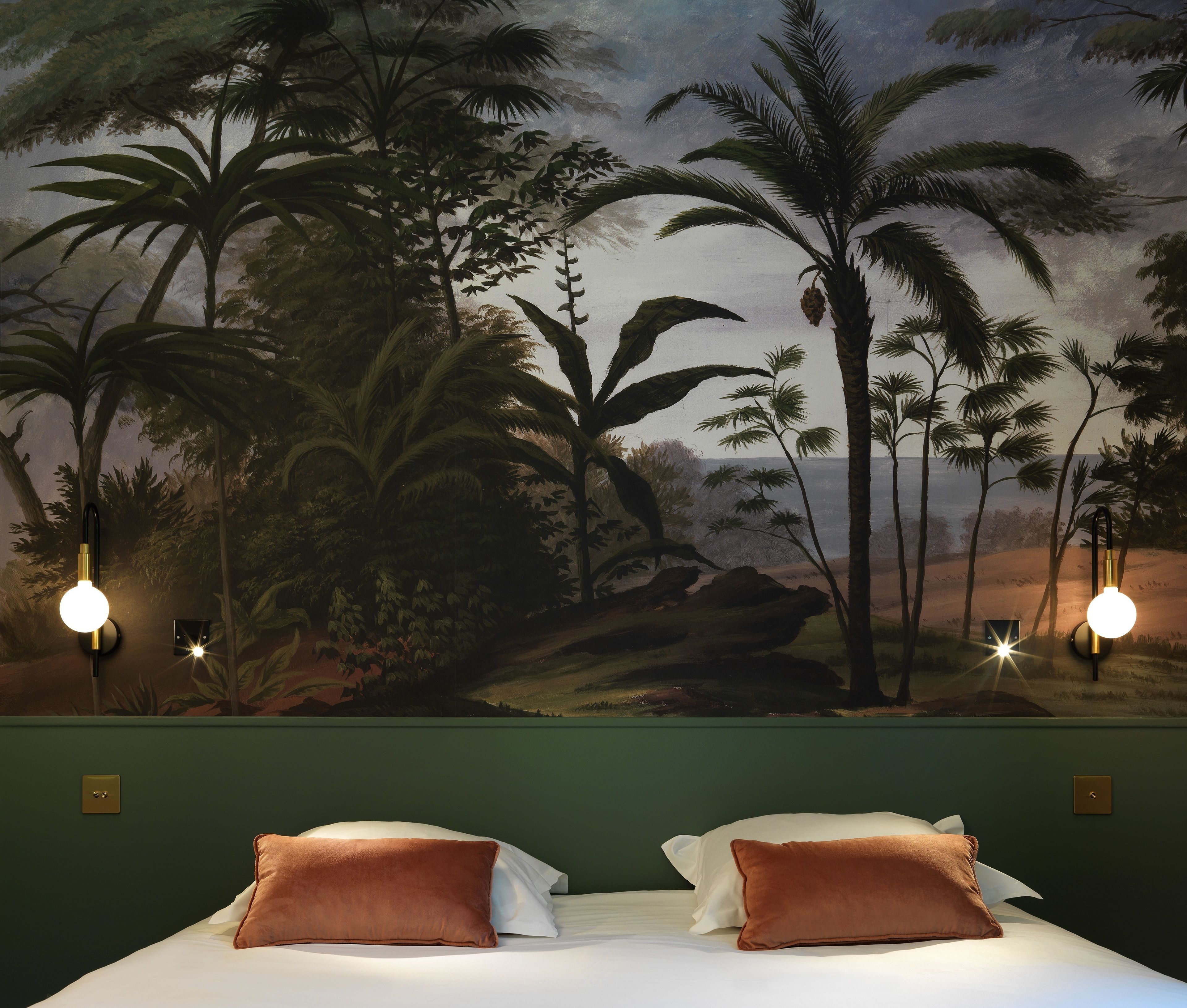 Une chambre à coucher ornée d’une fresque représentant un paysage tropical, avec deux lampes de chevet et un lit avec deux oreillers.