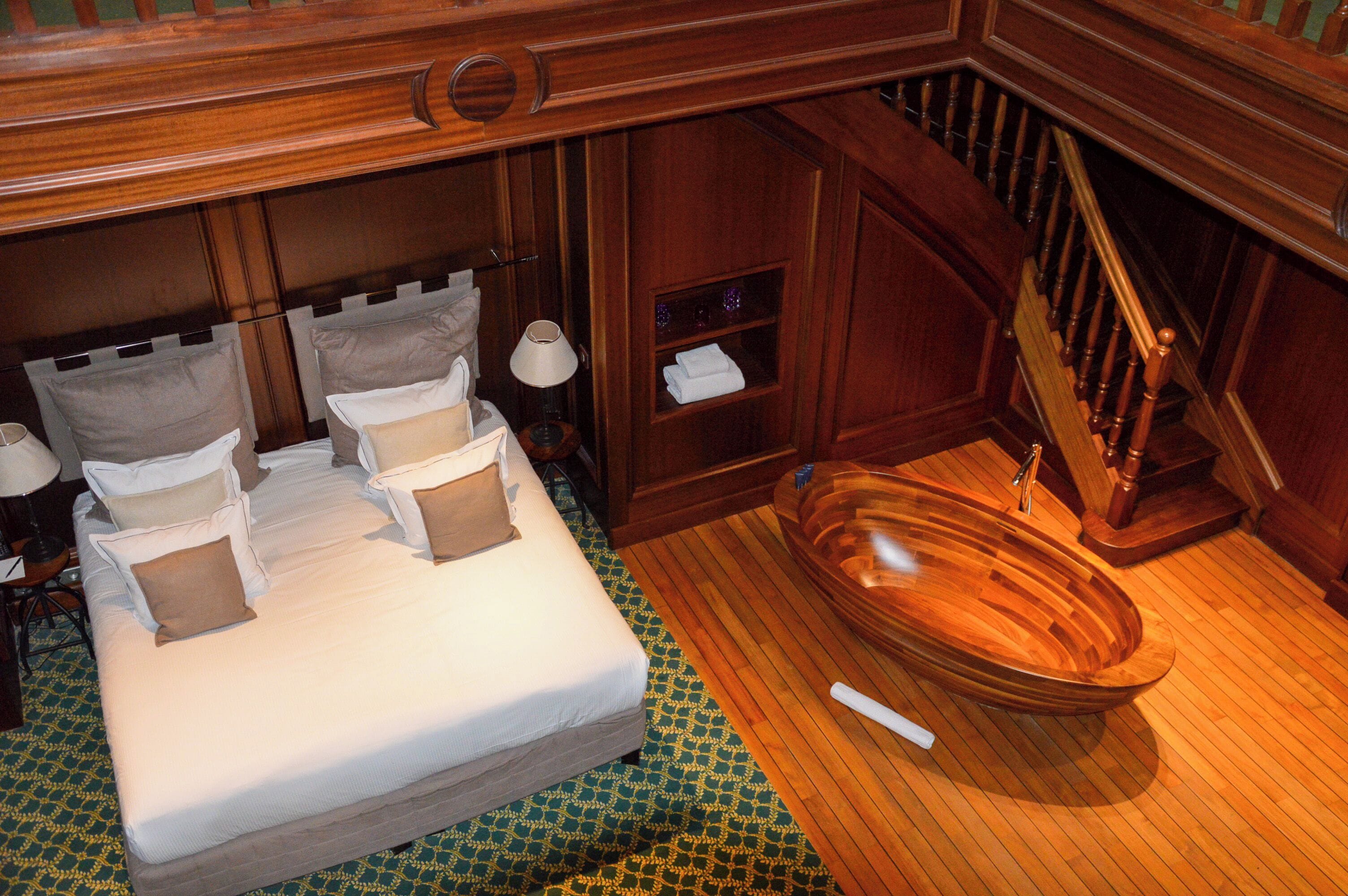 A wooden-staircase leading to a bathroom with a wooden bathtub, a bed with pillows, and a patterned floor.