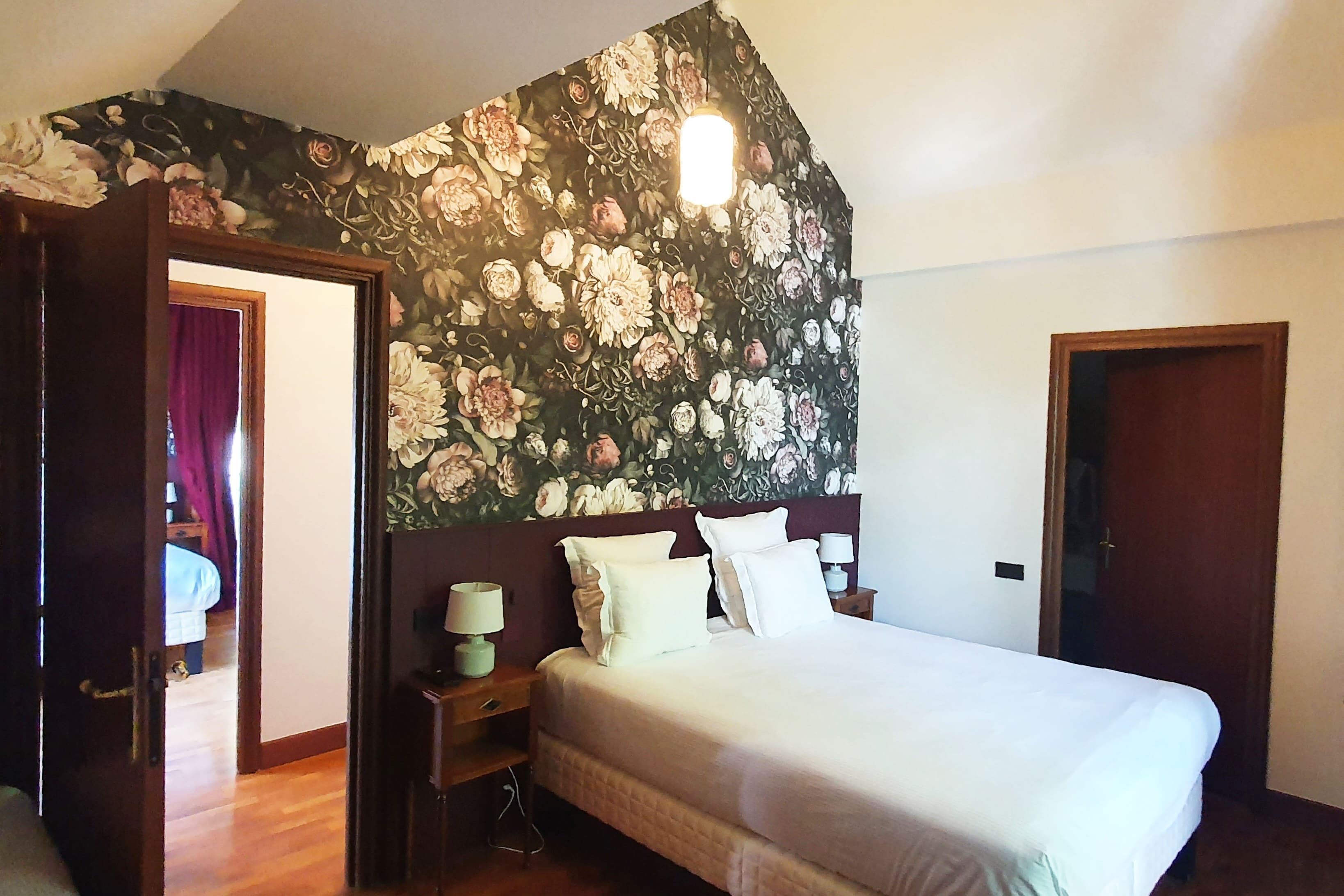 A bedroom with a floral wallpaper, a bed with white linens, a nightstand with a lamp, and a wooden door.
