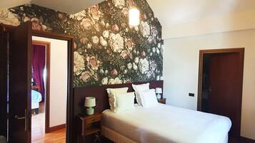 A bedroom with a floral wallpaper, a bed with white linens, a nightstand with a lamp, and a wooden door.
