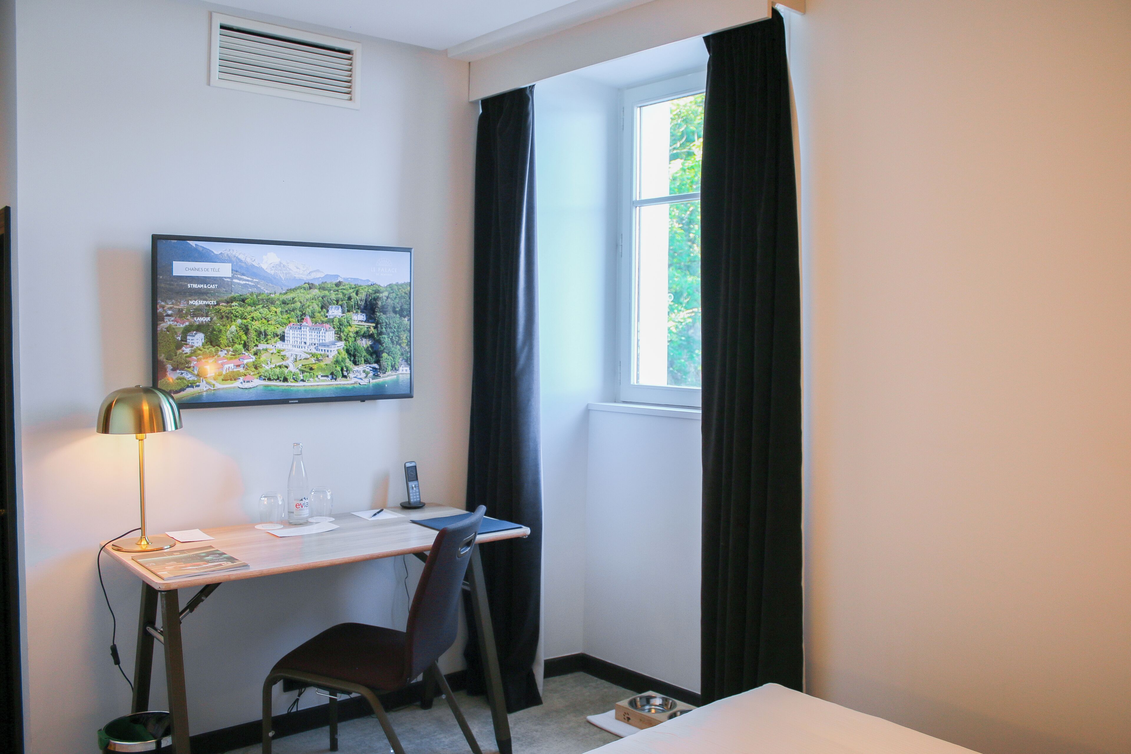 Une chambre d’hôtel équipée d’un bureau, d’une chaise, d’un lit, d’une fenêtre avec des rideaux, d’un tableau accroché au mur et d’un radiateur.