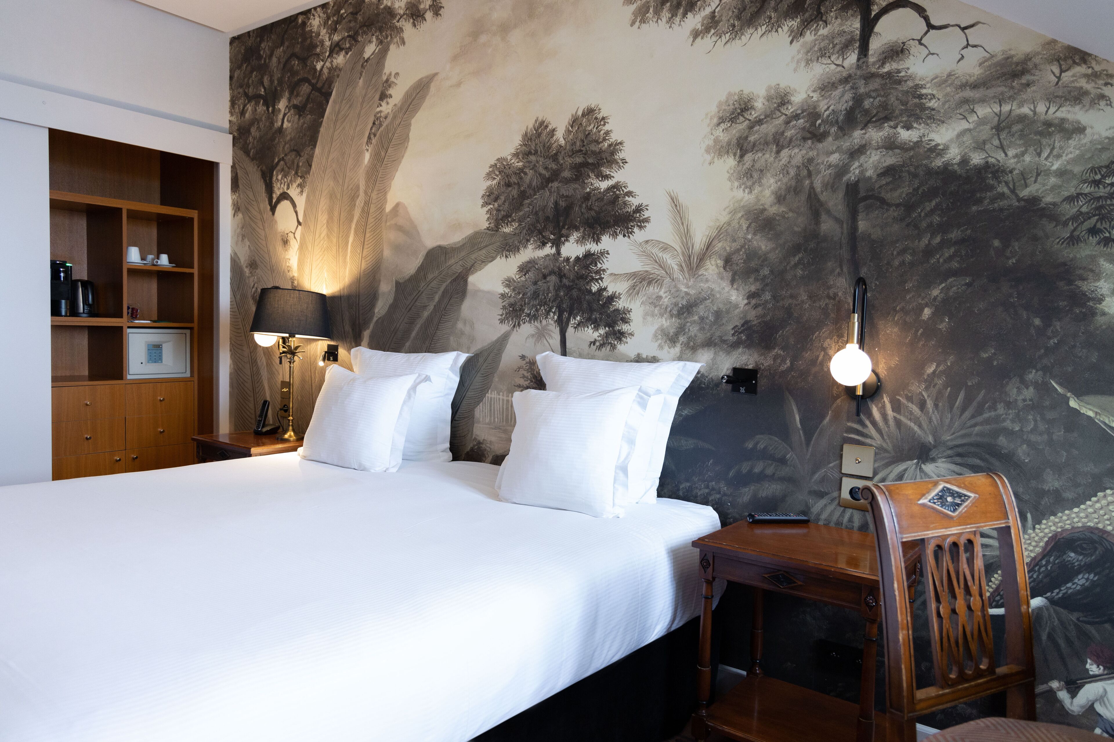 A hotel room with a large bed, a wooden nightstand with a lamp, a wooden wardrobe, and a mural of trees on the wall.