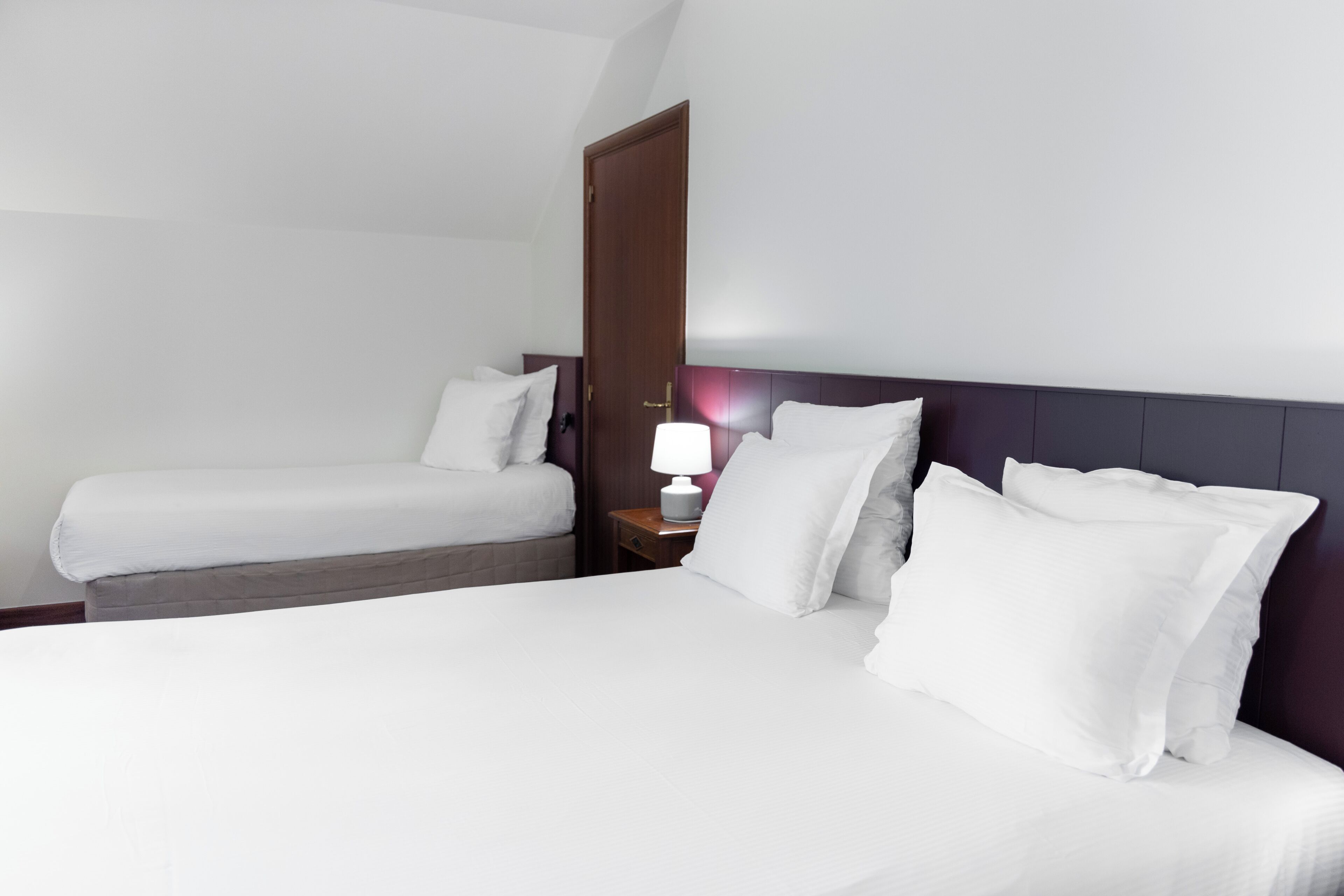 A hotel room with a large bed, a smaller bed, a nightstand with a lamp, and a door.