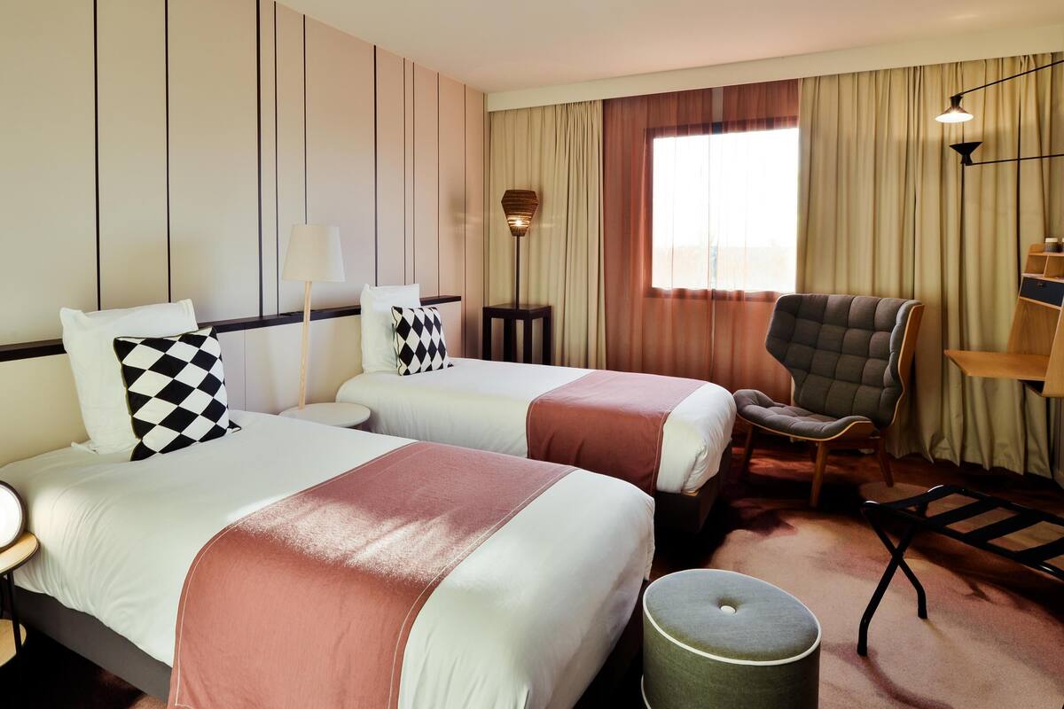 A hotel room with two beds, a chair, a lamp, and a small table.