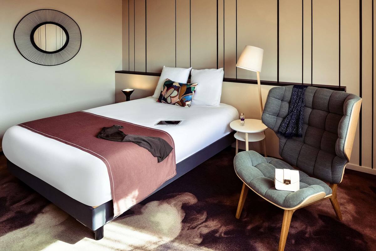 A hotel room with a bed, a chair, a bedside table, a lamp, and a wall-mounted mirror.
