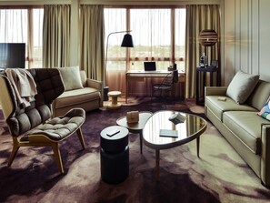 Una sala de estar moderna con sofá, sillón y mesa de centro, un escritorio con silla, y un ventanal con cortinas.