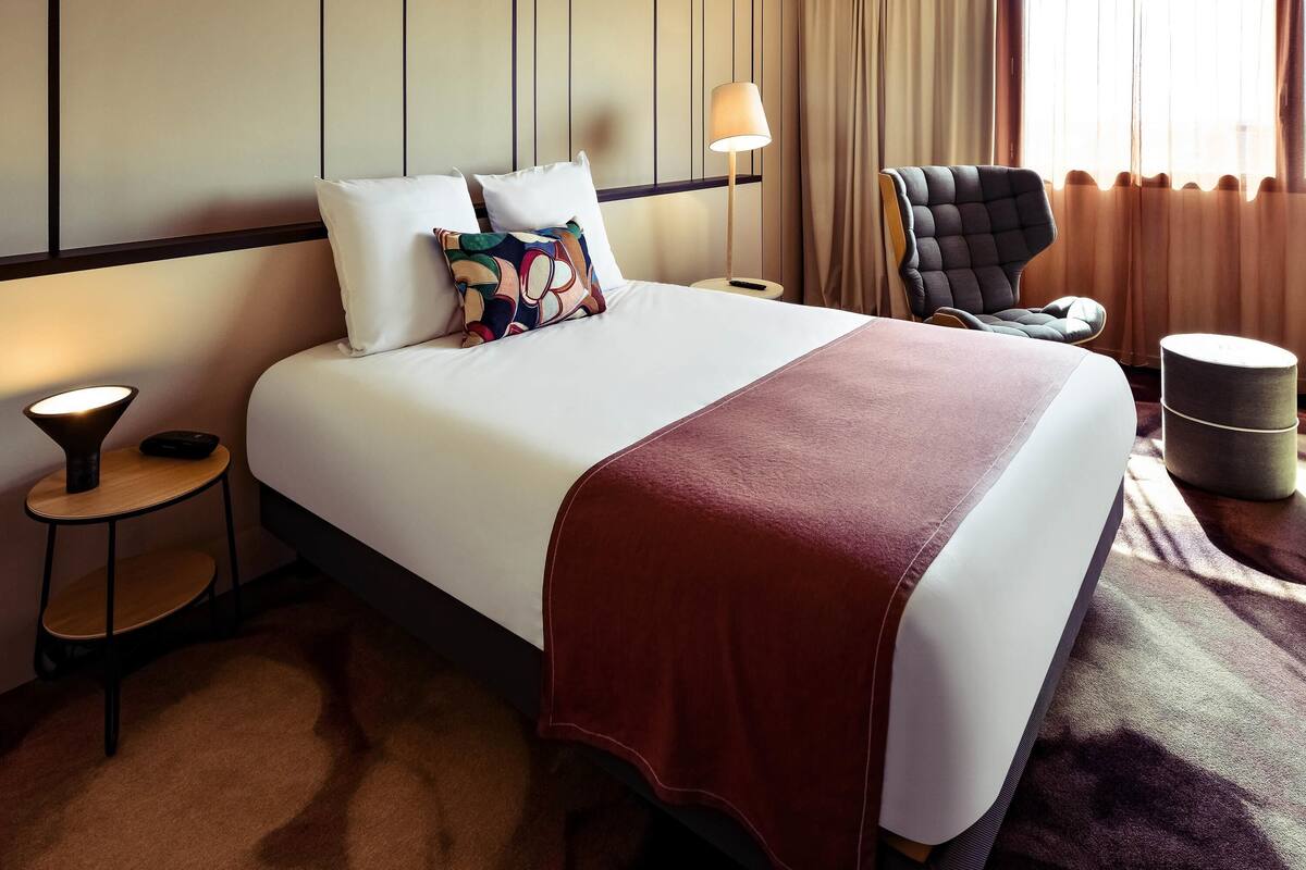 A hotel room with a large bed, a chair, a small table, and a lamp.