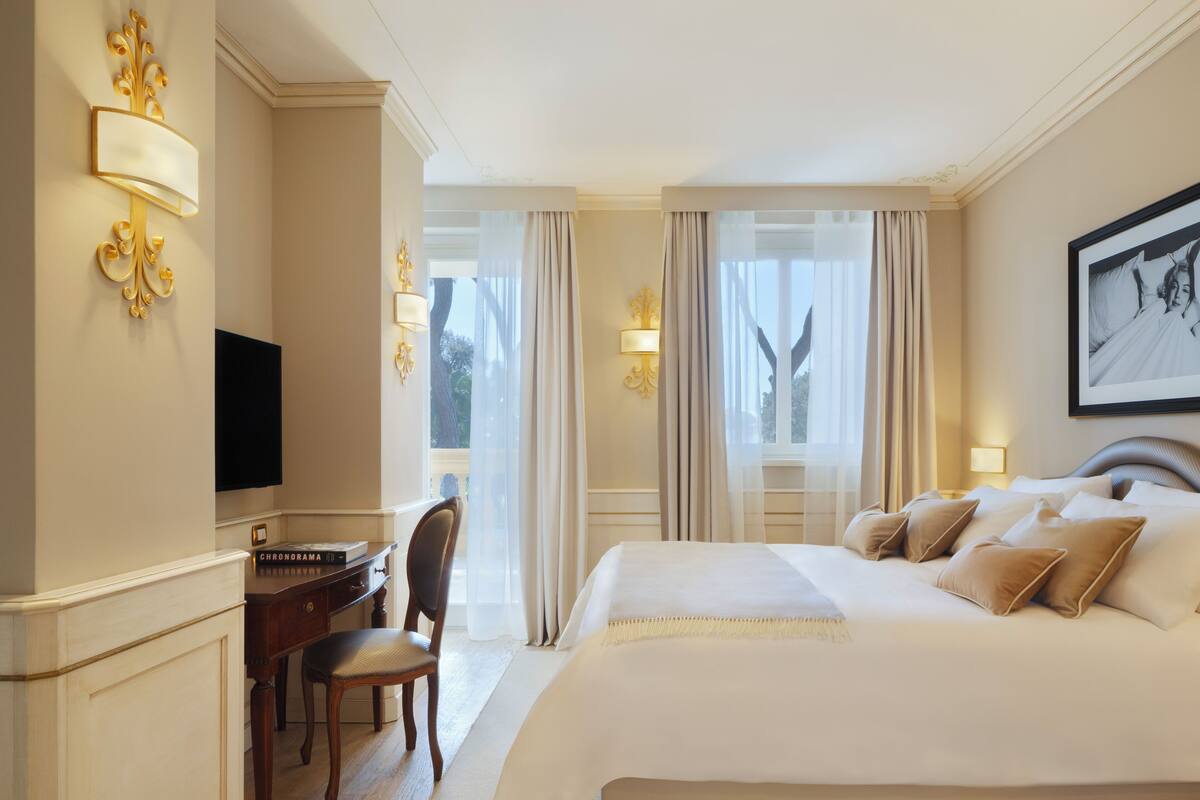 A hotel room with a large bed, a desk with a chair, a television, and a window with curtains.