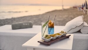 Ein Tisch mit einer Auswahl an Snacks und Getränken, einem Kissen und einer Decke am Strand mit Sonnenuntergang.