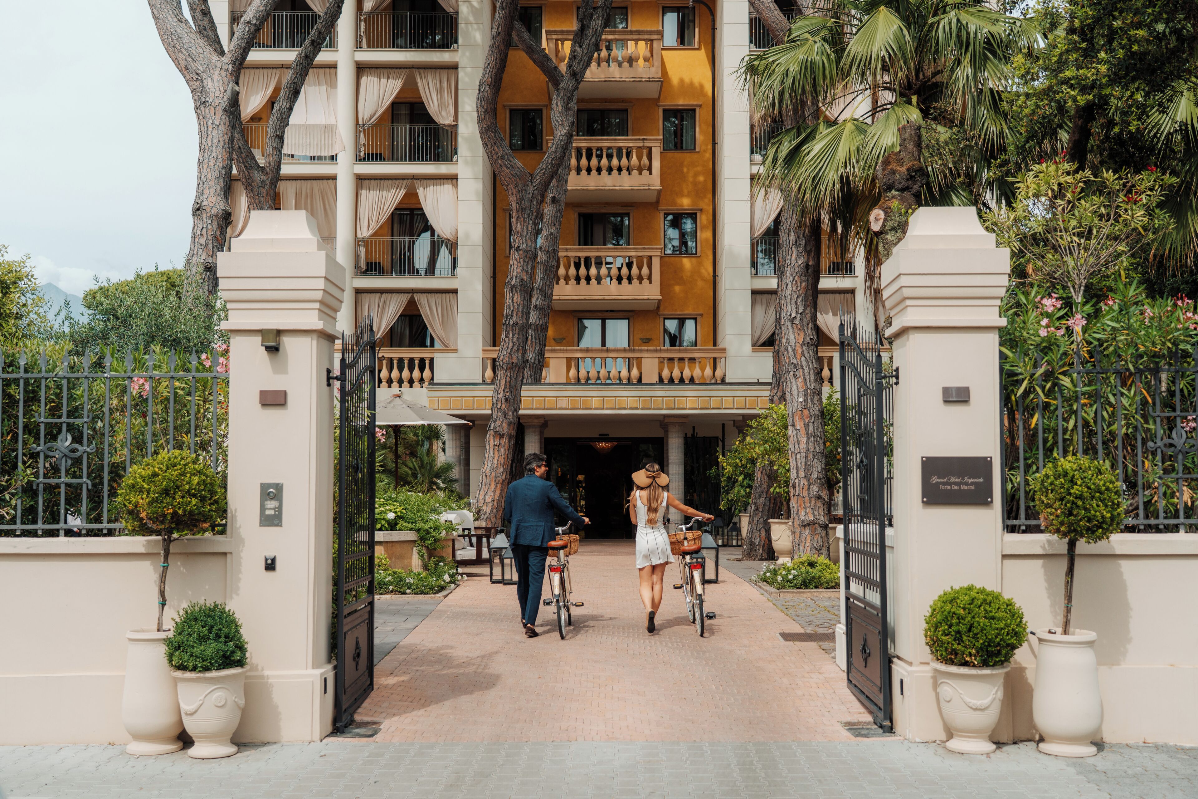 A gated entrance to a hotel with a central pathway, surrounded by potted plants and trees.