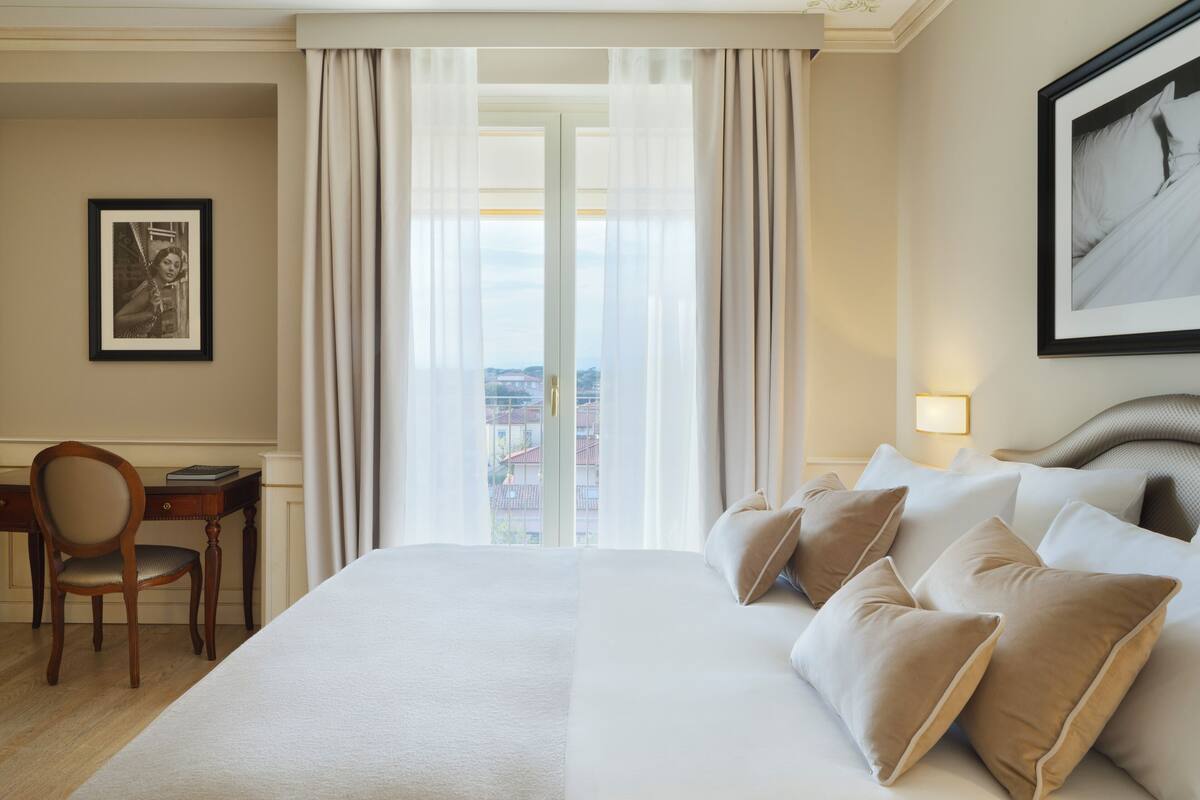 A hotel room with a large bed, a desk with a chair, a framed picture on the wall, and a window with curtains.