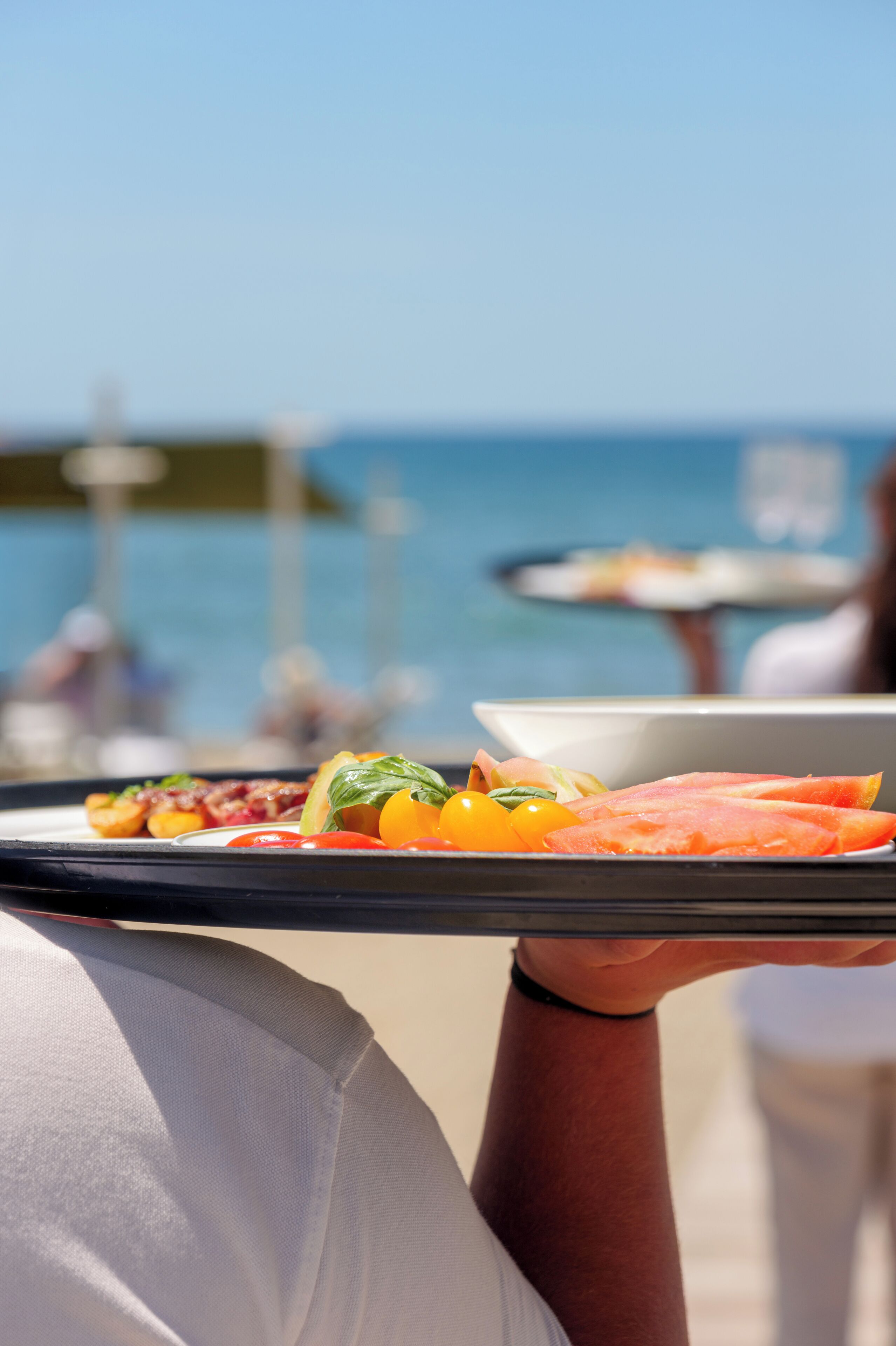 A person holding a tray with food items, including tomatoes and basil, with a beach and ocean in the background.