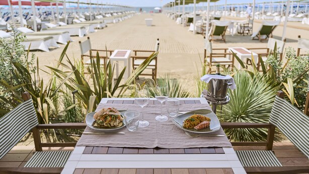 Ein Essbereich direkt am Strand mit Tischen und Stühlen, Blick auf den Strand sowie ein Tisch mit Speisen und Gläsern.
