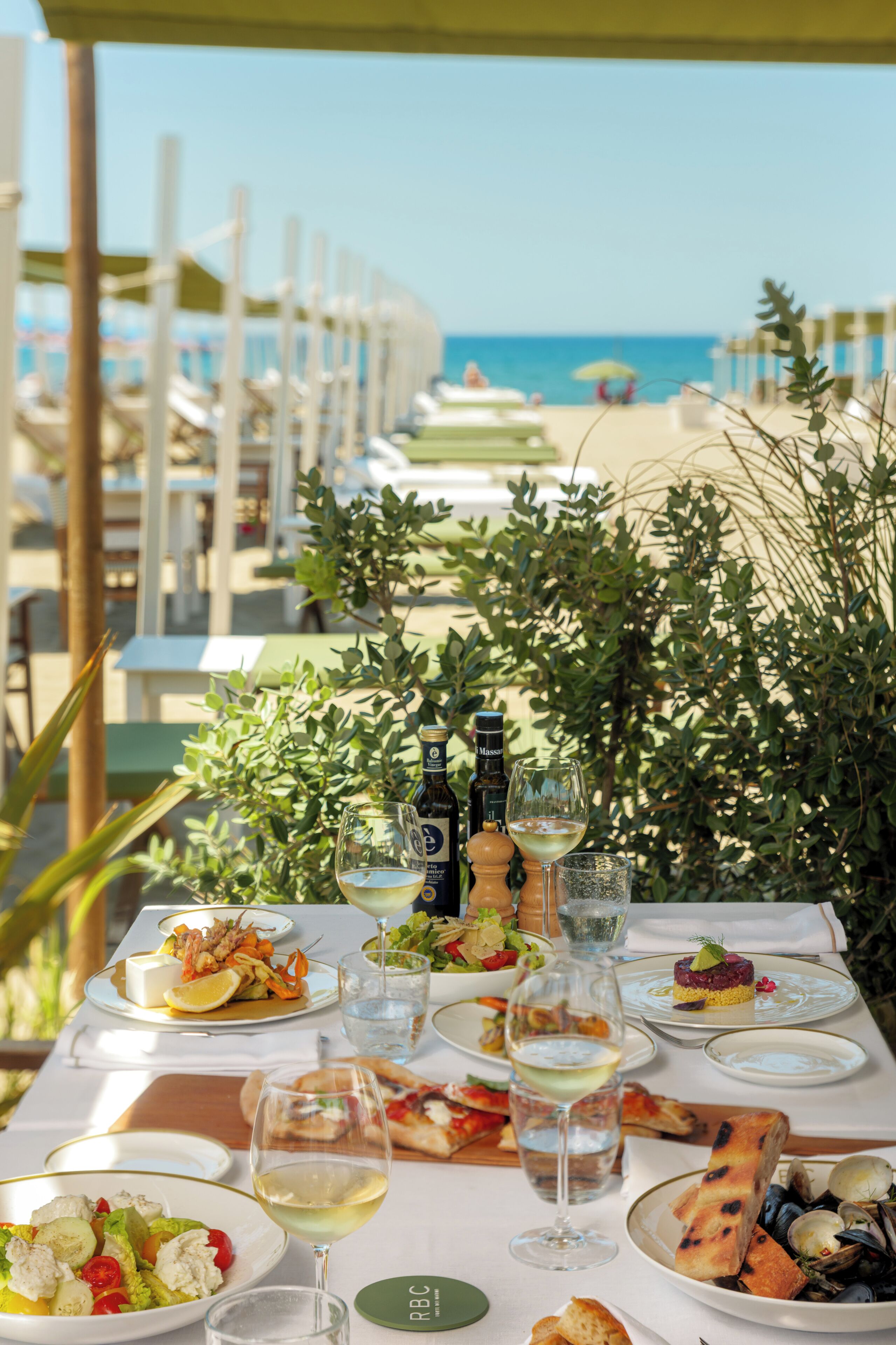 A table set for dining with various dishes, glasses of wine, and a bottle of olive oil, overlooking a beach.