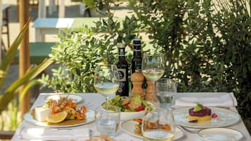 A table set for dining with various dishes, glasses of wine, and a bottle of olive oil, overlooking a beach.
