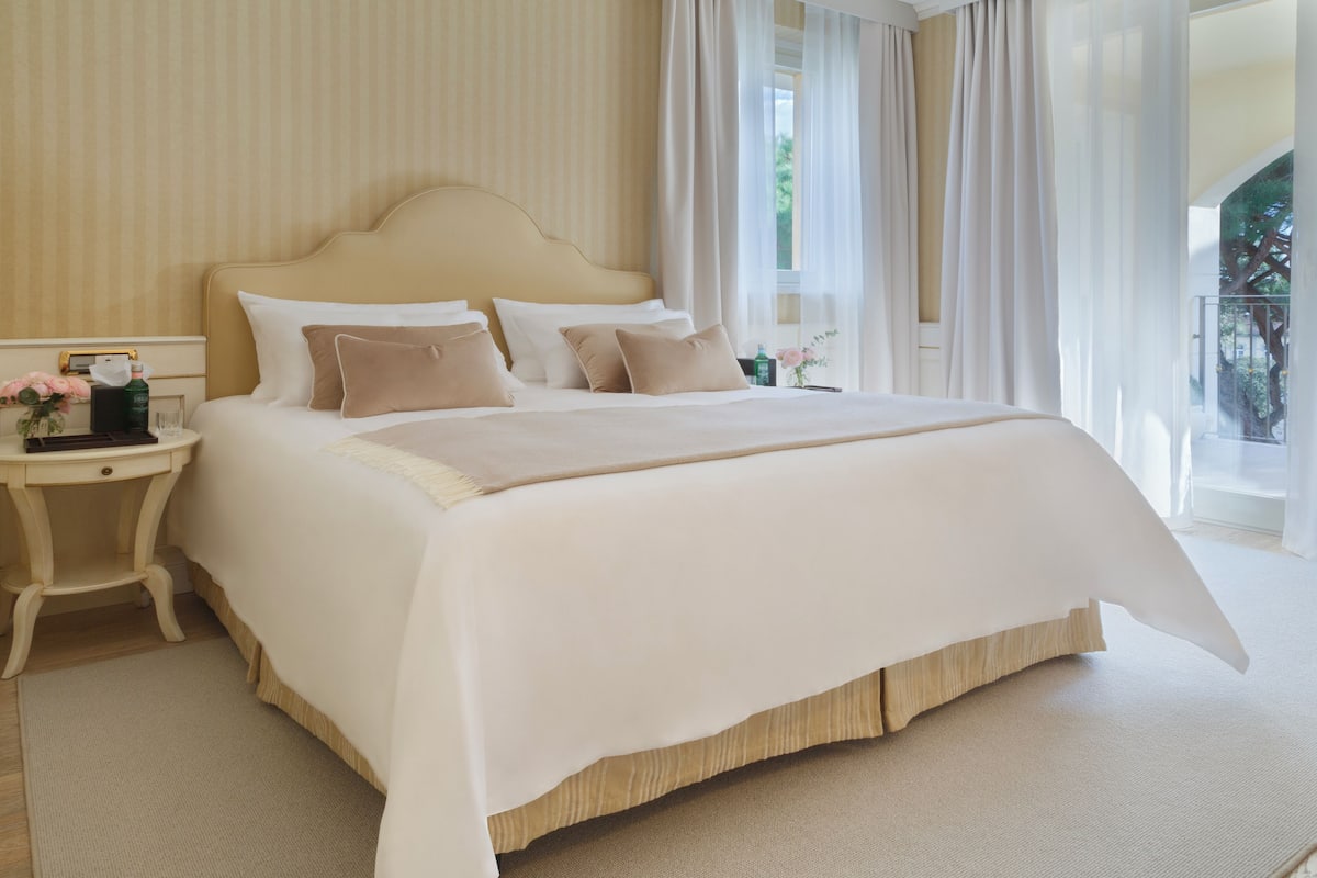 A large bed with a wooden headboard, a bedside table with a lamp, and a view of the outdoors through sheer curtains.