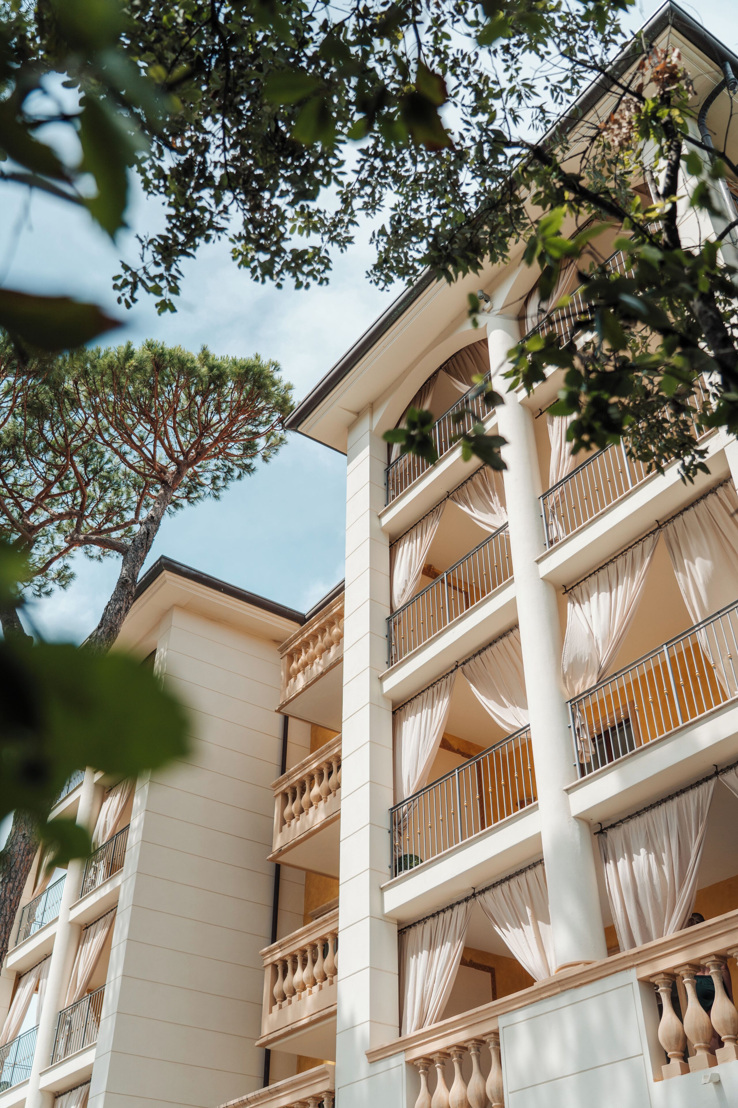 A multi-story building with balconies, white walls, and a tree in the foreground.