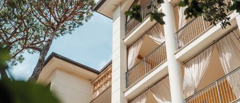 A multi-story building with balconies, white walls, and a tree in the foreground.