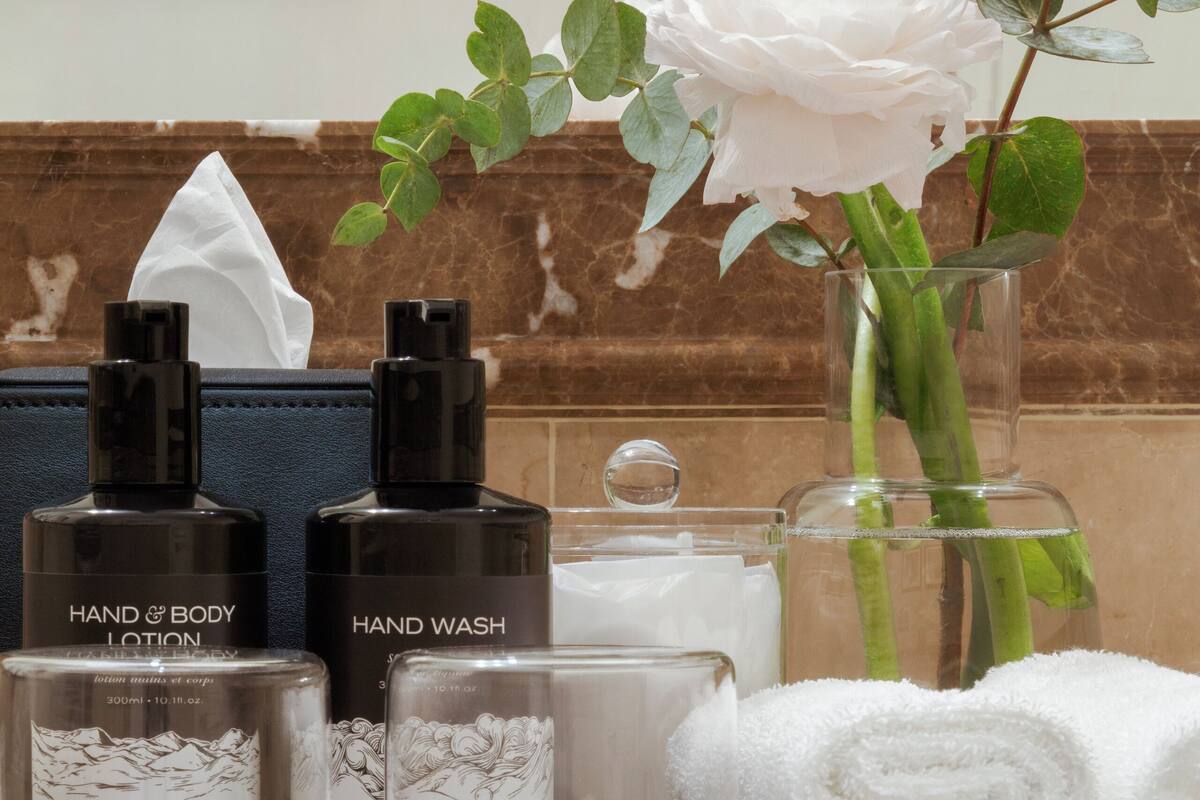 A bathroom vanity with hand and body lotion, hand wash, a rolled white towel, and a vase with a white rose.