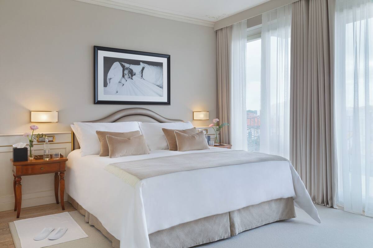 A hotel room with a large bed, two bedside tables with lamps, a framed picture on the wall, and a view of the city through the window.