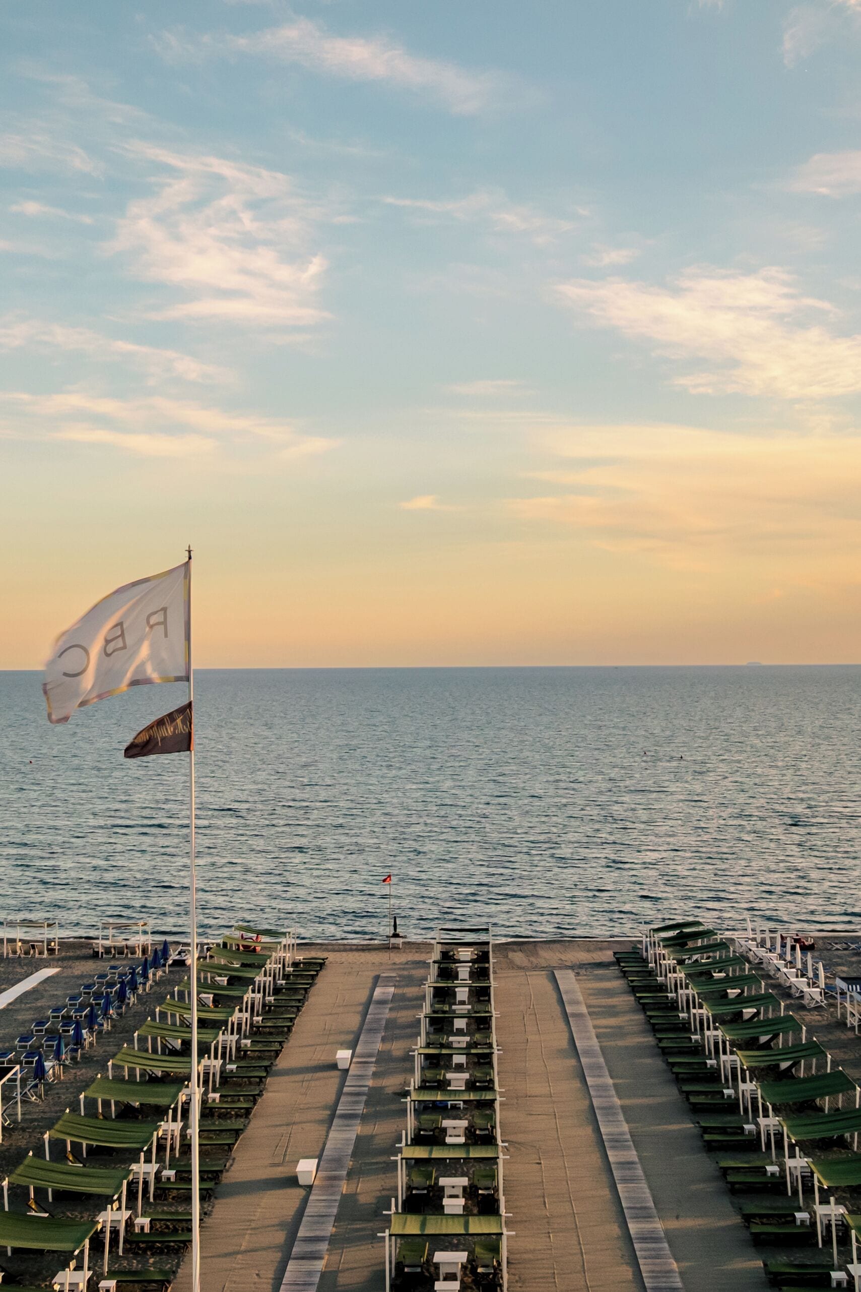 A pier with rows of beach chairs and umbrellas, a flag, and a view of the sea.