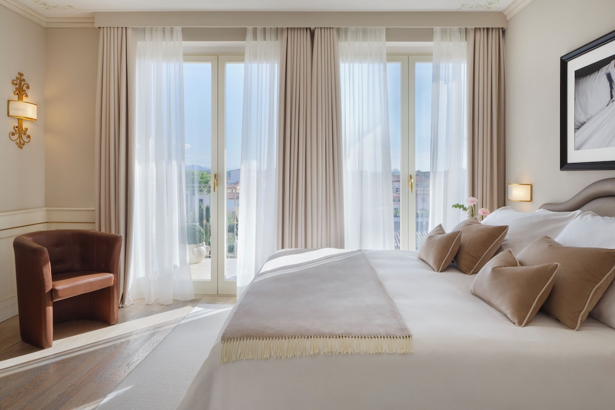 A hotel room with a large bed, a chair, a view through the window, and a framed picture on the wall.