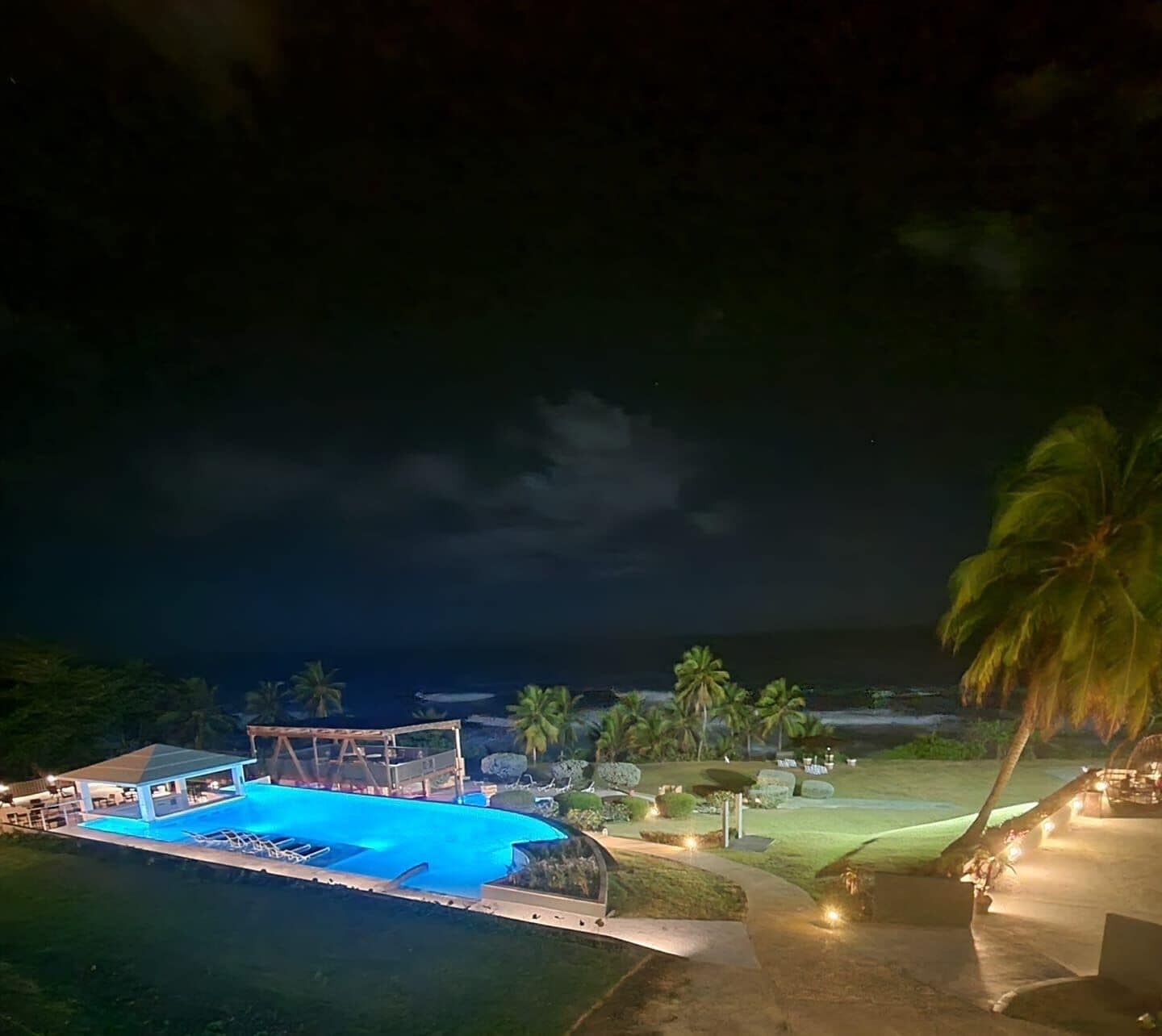 A nighttime view of a resort with a swimming pool, palm trees, and a view of the ocean.