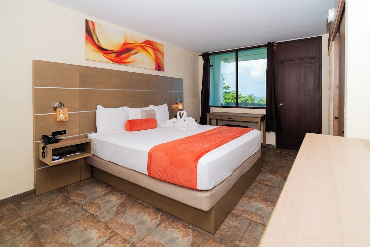 A hotel room with a bed, bedside table, lamp, artwork, window, and a view of greenery.