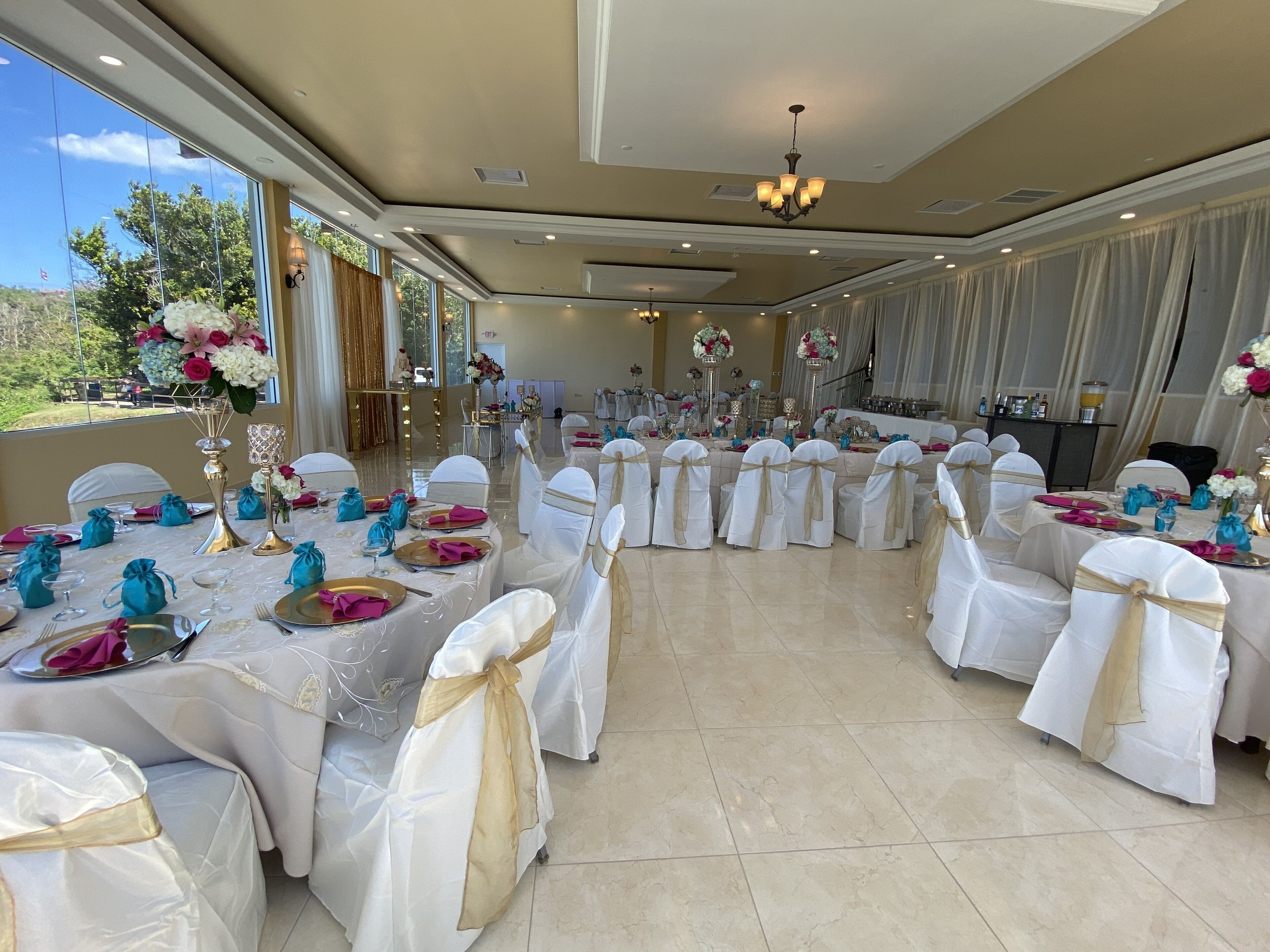 Un salón de banquetes con mesas redondas dispuestas para un evento formal, con manteles blancos, fajos dorados y servilletas color turquesa.