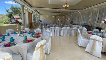Un salón de banquetes con mesas redondas dispuestas para un evento formal, con manteles blancos, fajos dorados y servilletas color turquesa.