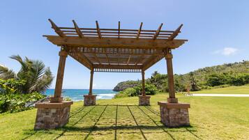 A wooden pergola structure with stone bases, set on a grassy area with a view of the ocean and hills in the background.