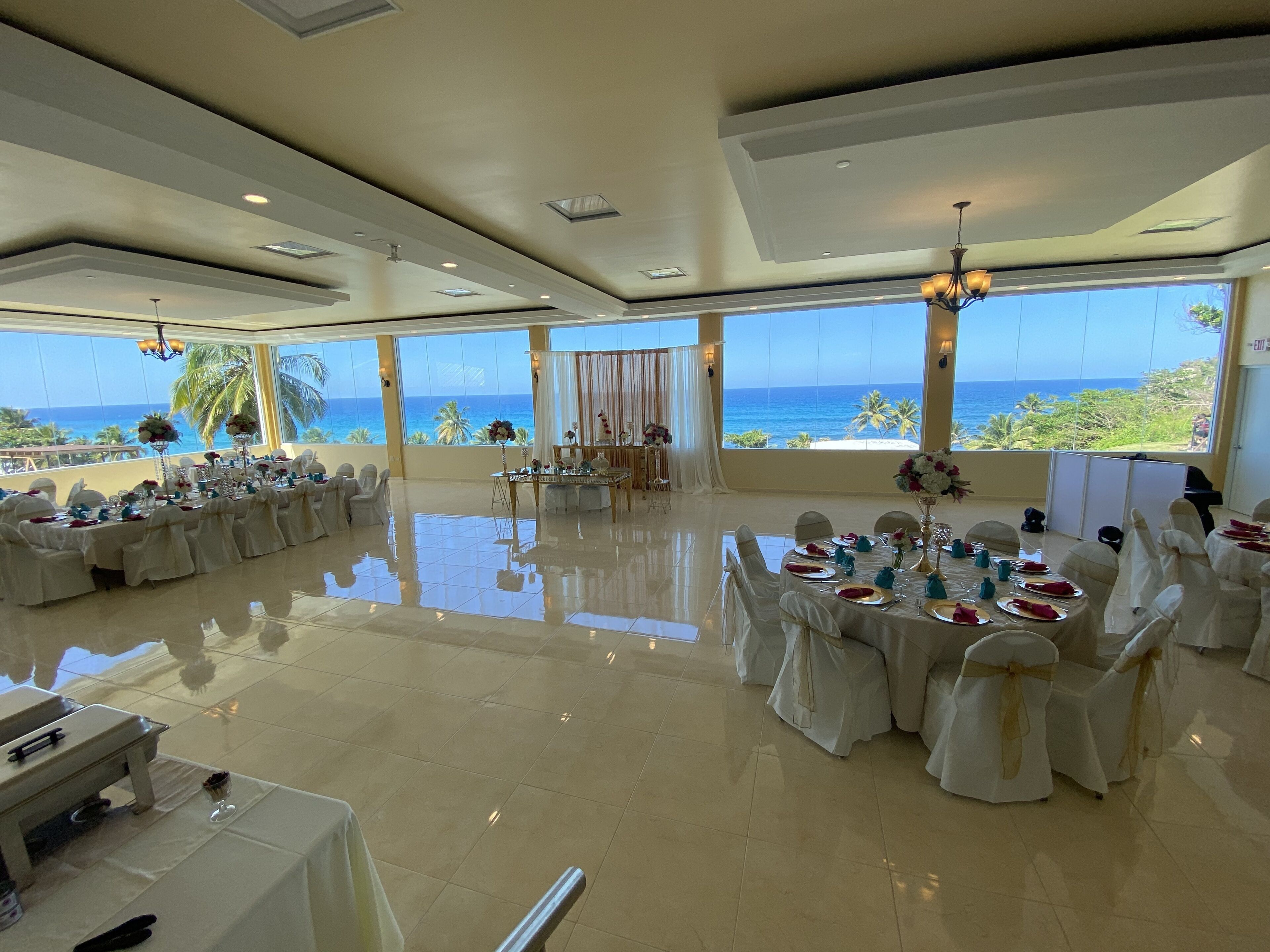 Un espacioso salón de banquetes con amplias ventanas que ofrecen vistas al océano, suelos pulidos y mesas dispuestas con mantelería blanca.