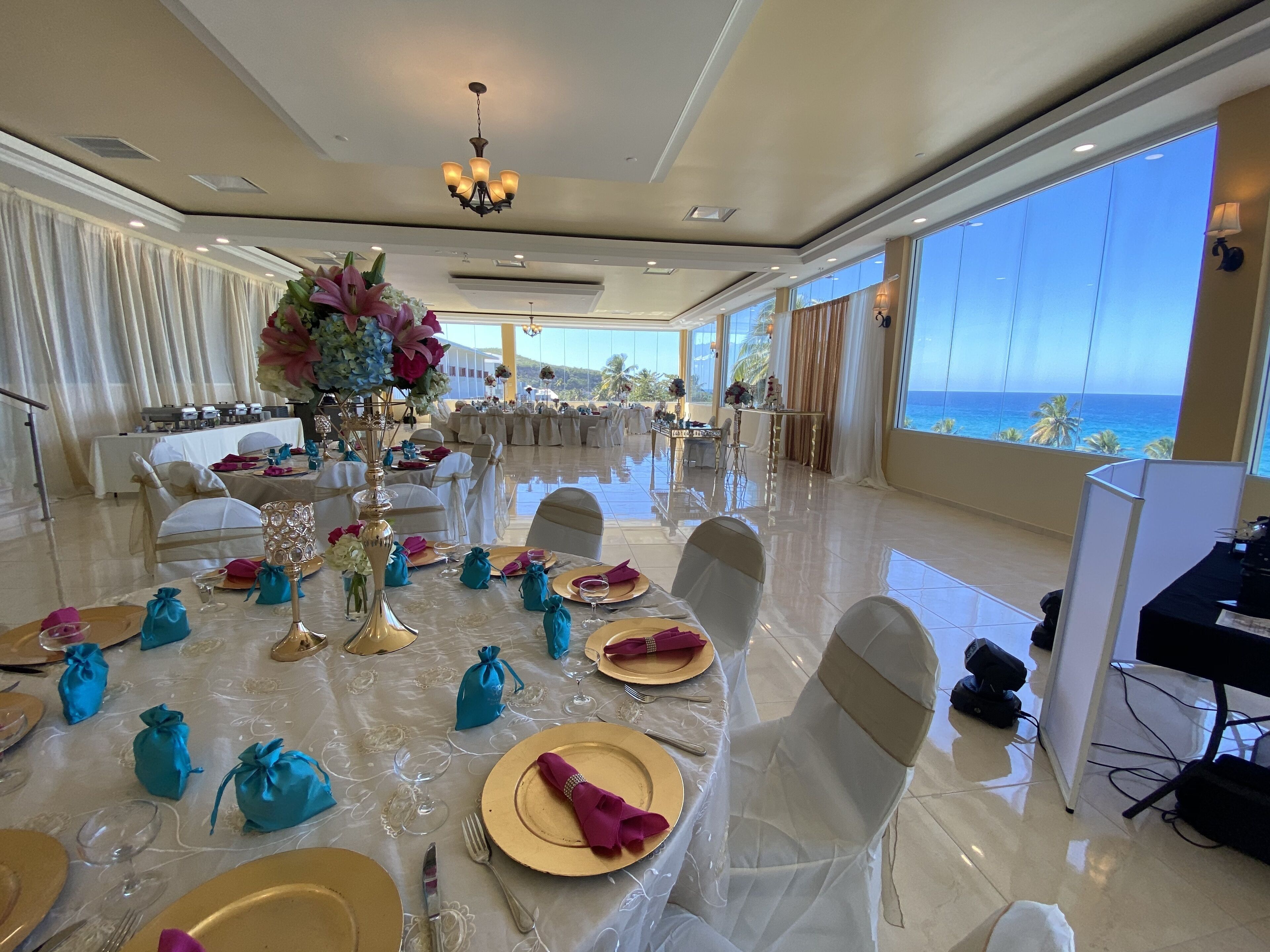 A spacious dining area with tables set for a formal event, large windows offering a view of the ocean, and a chandelier hanging from the ceiling.