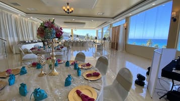 A spacious dining area with tables set for a formal event, large windows offering a view of the ocean, and a chandelier hanging from the ceiling.