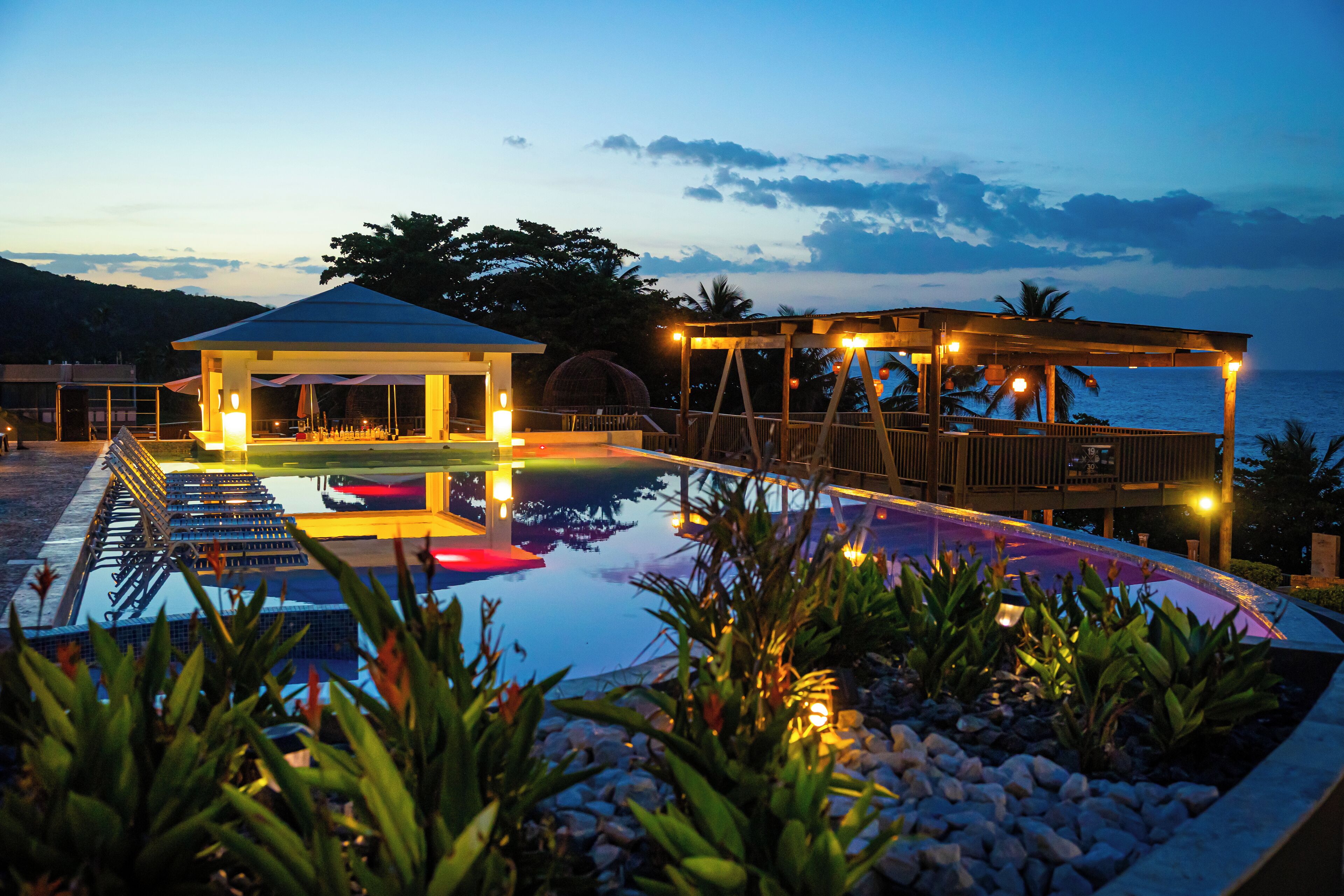 A pool area with a covered seating area and a view of the sea at dusk.
