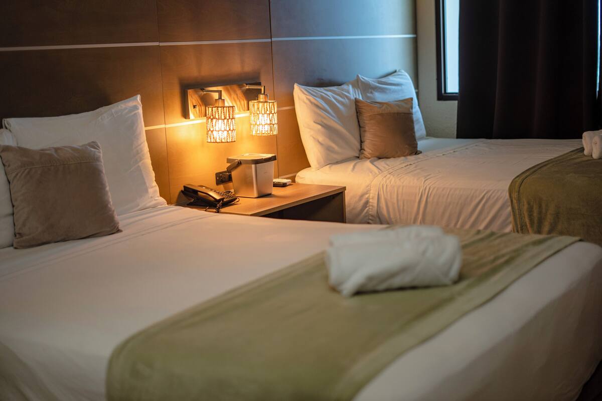 A hotel room with two beds, a nightstand with a phone and lamp, and a window with curtains.