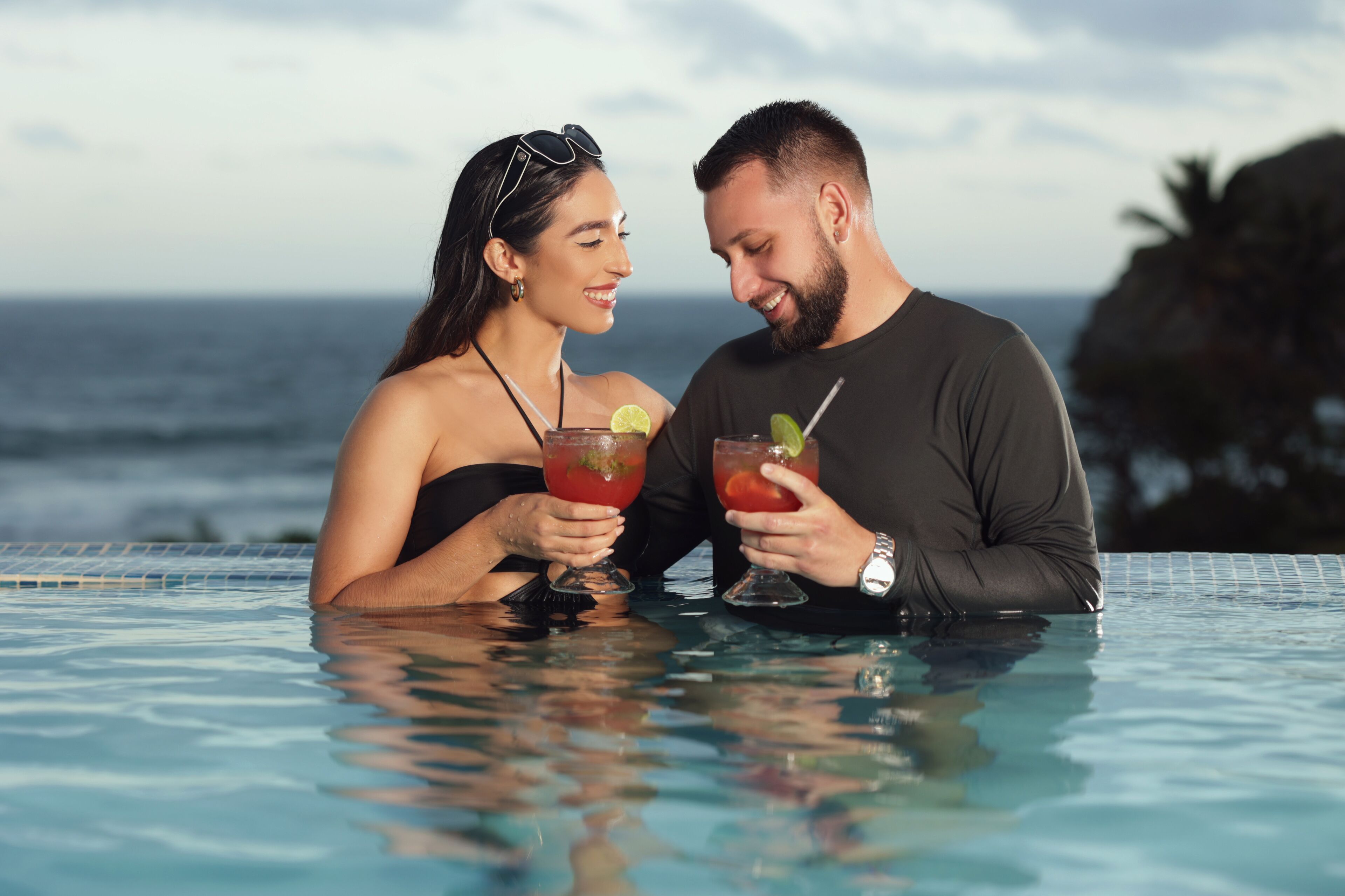 A couple in a swimming pool with drinks, overlooking the ocean.